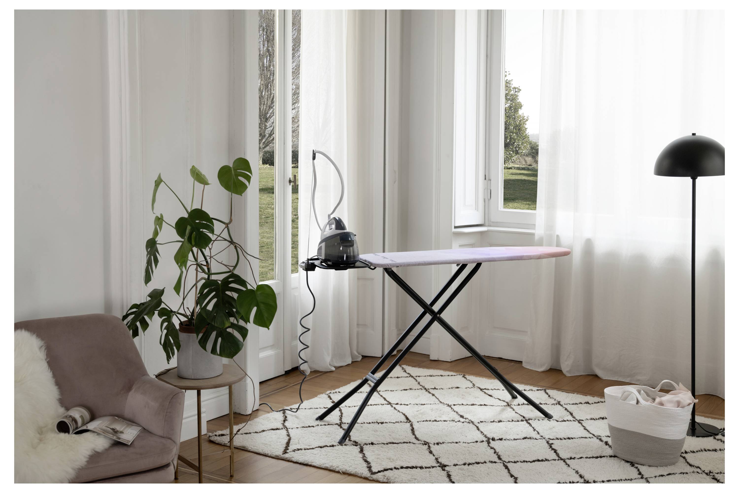 Helles Wohnzimmer mit einem kompakten Bügelbrett am Fenster, neben einer Stehlampe, einer Topfpflanze, einem Stuhl und einem Wäschekorb.
