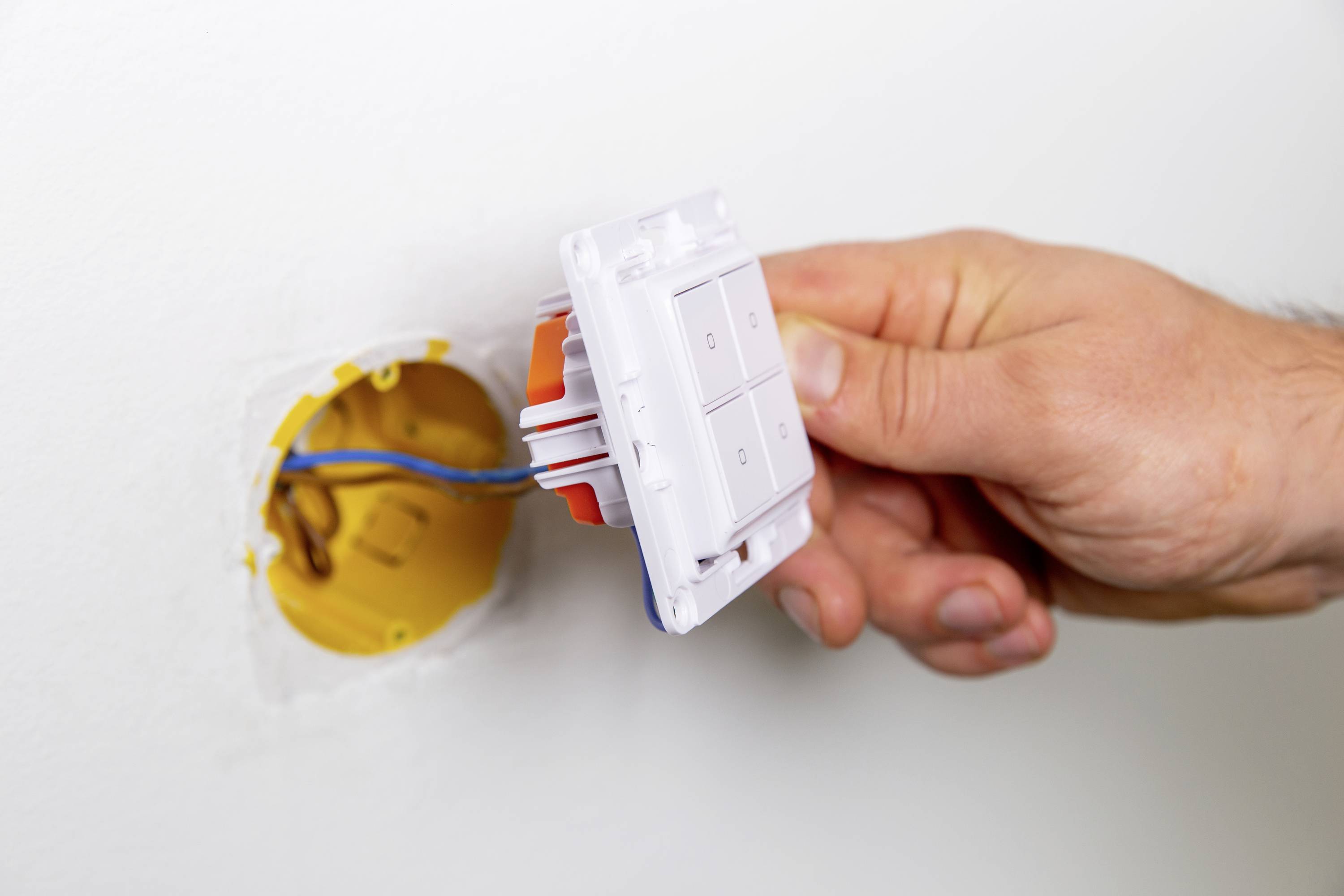 Eine Hand installiert oder entfernt einen Lichtschalter aus einer Wandöffnung. Elektrische Drähte sind sichtbar.