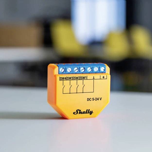 Ein kleines orangefarbenes Gerät mit blauen Anschlüssen und Aufschrift 'Shelly DC 5-24 V' auf einem Tisch, unscharfer Hintergrund.