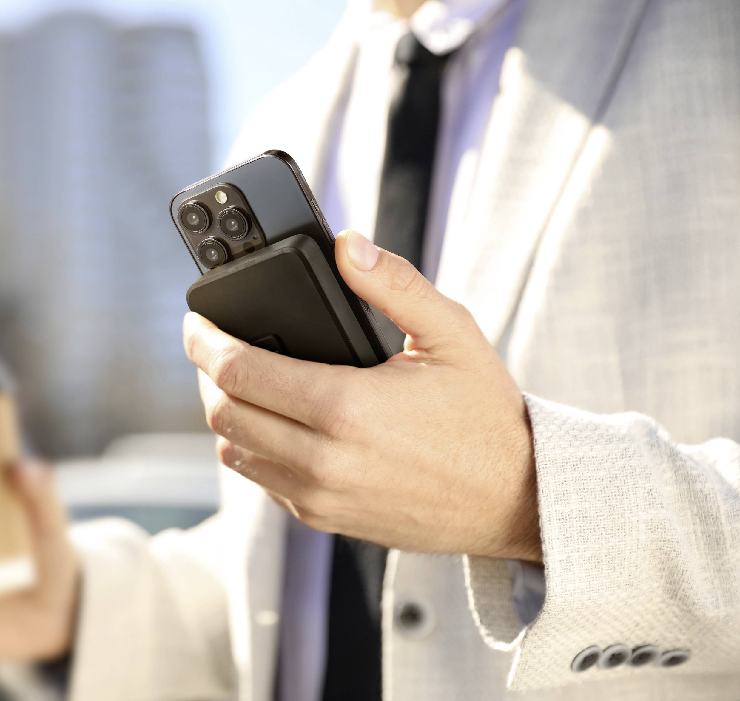Ein Mann in einem hellen Anzug hält ein Smartphone und einen Kaffeebecher. Im Hintergrund sind unscharfe Gebäude zu sehen.