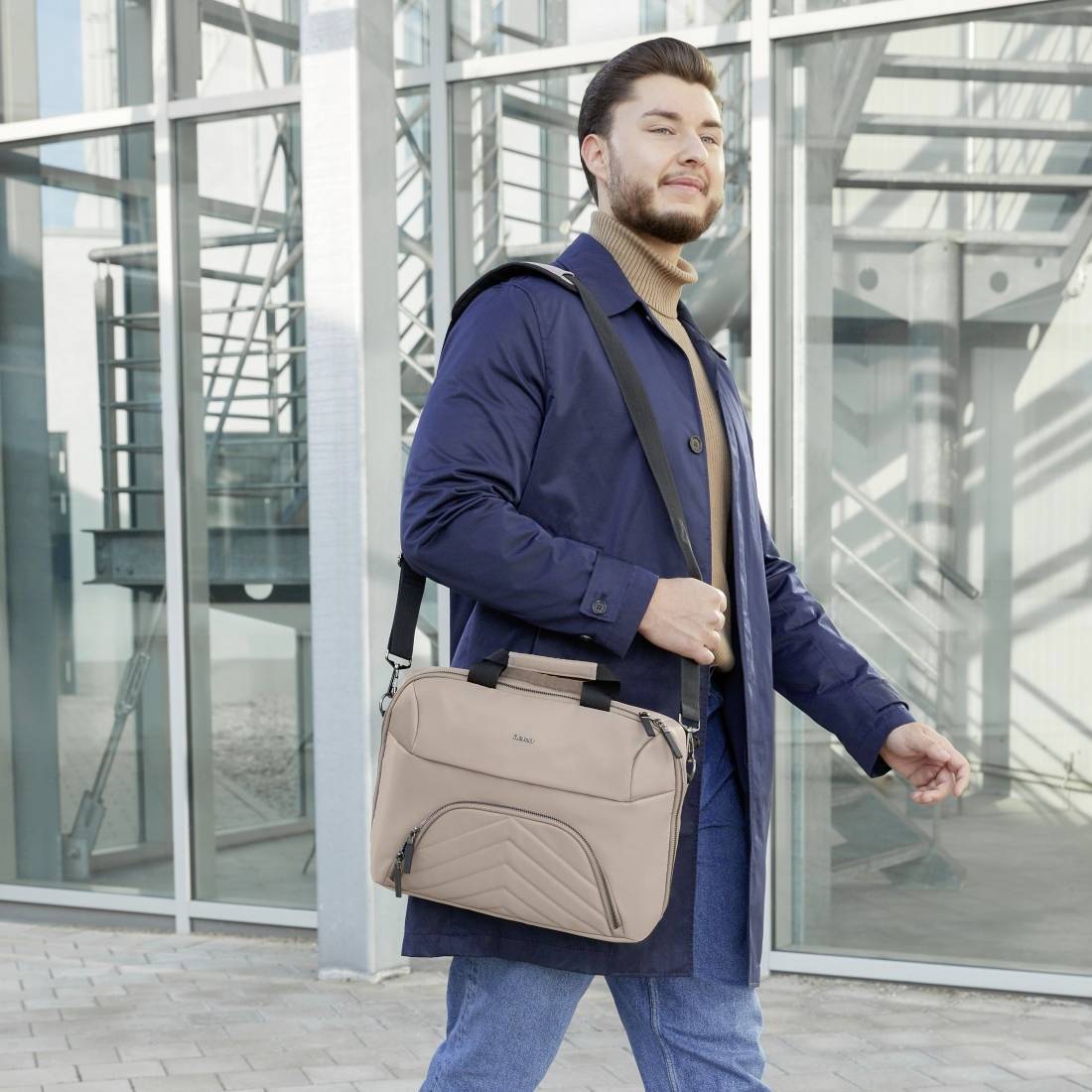 Ein Mann im blauen Mantel und Jeans steht selbstbewusst vor einem modernen Gebäude und trägt eine beigefarbene Umhängetasche.