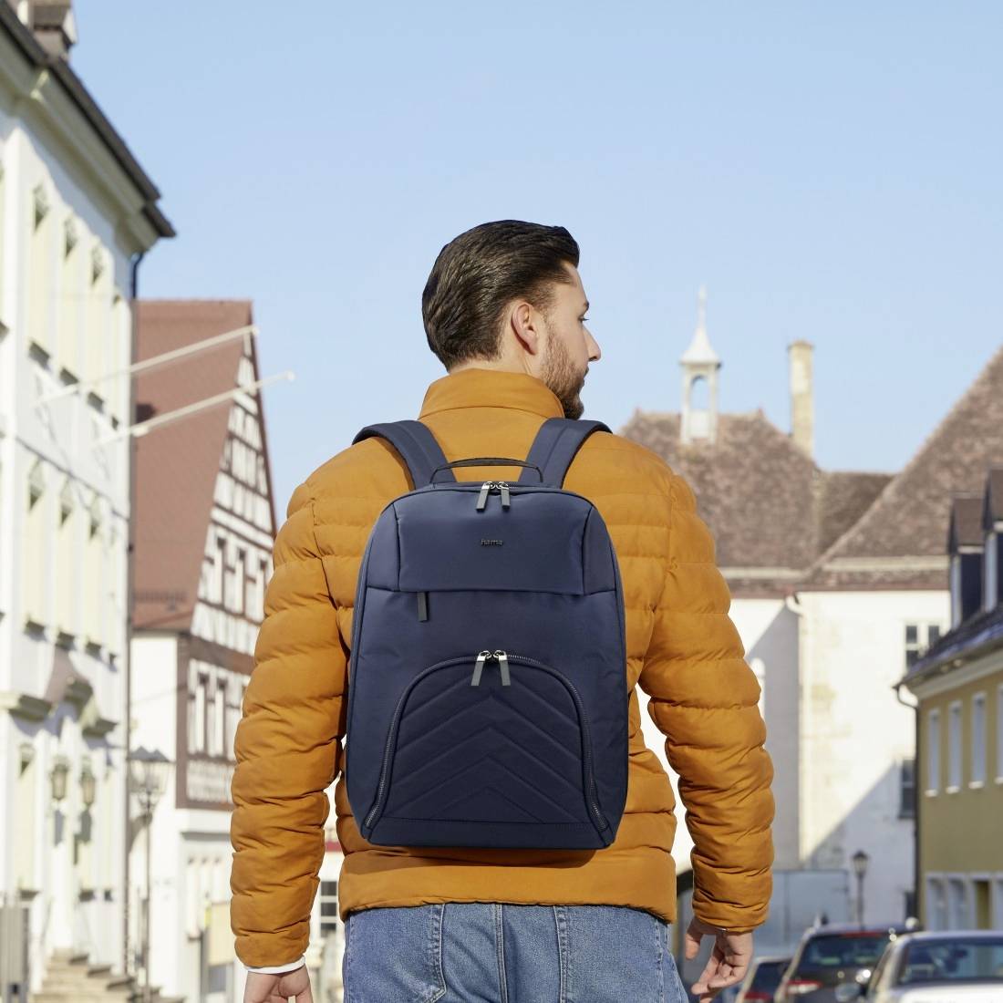 Ein Mann in blauer Jeans und orangefarbener Jacke mit Rucksack geht eine europäische Straße entlang. Historische Gebäude im Hintergrund.