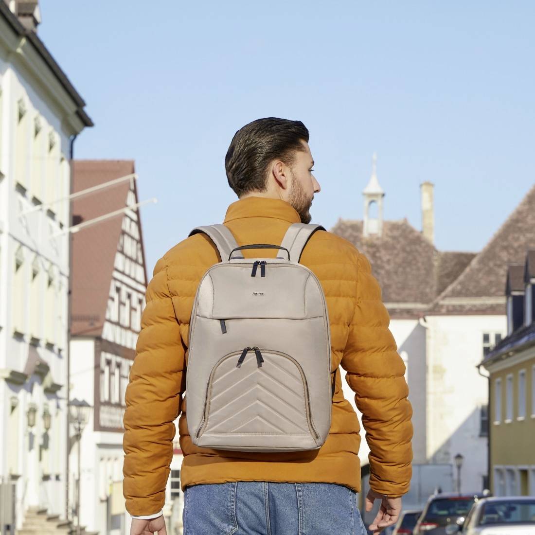 Eine Person mit brauner Jacke und grauem Rucksack steht auf einer sonnigen, europäischen Straße mit historischen Gebäuden.