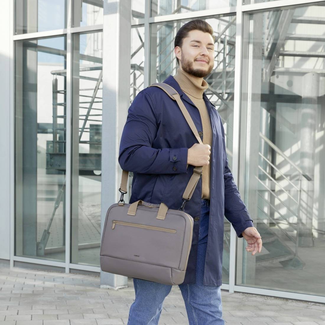 Ein Mensch geht mit einer Bürotasche in der Hand und trägt einen blauen Mantel. Im Hintergrund ist eine moderne Glasfassade sichtbar.