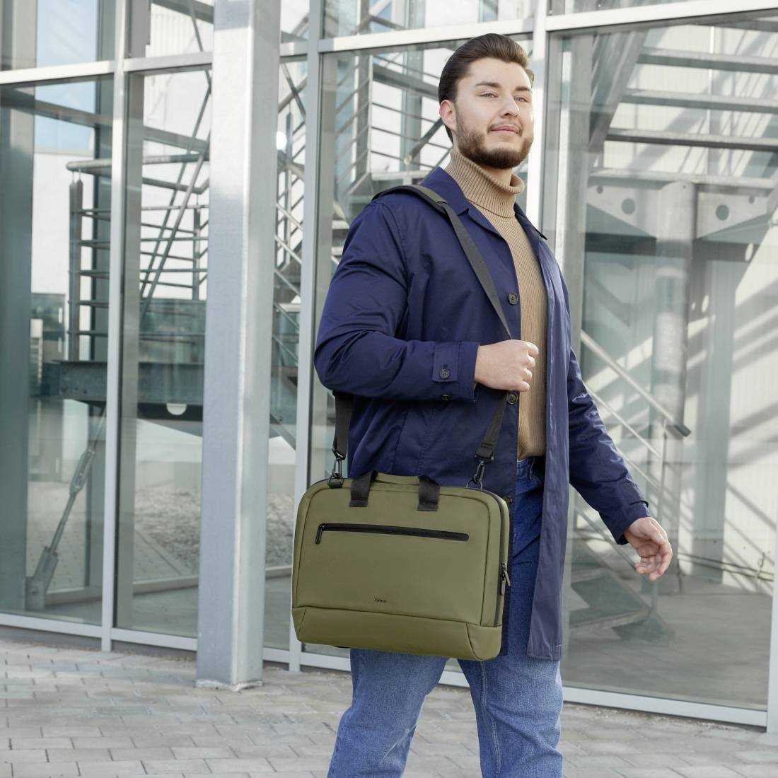 Ein Mann steht vor einem modernen Gebäude mit Glasfassade. Er trägt einen Mantel, hält eine olivgrüne Tasche und schaut selbstbewusst.