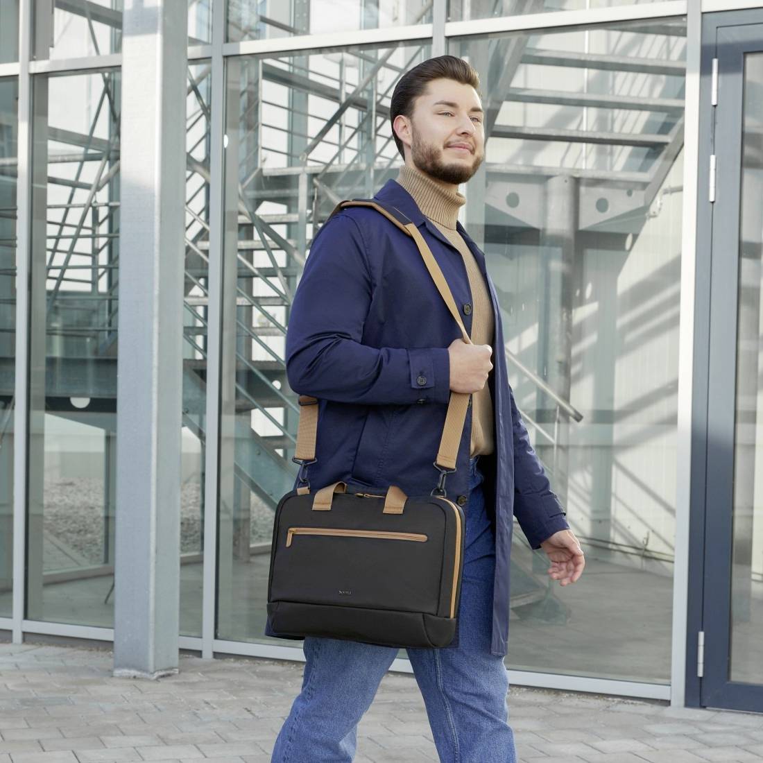 Ein Mann steht draußen vor einem modernen Gebäude, trägt eine dunkelblaue Jacke, einen braunen Rollkragenpullover und hält eine schwarze Aktentasche.