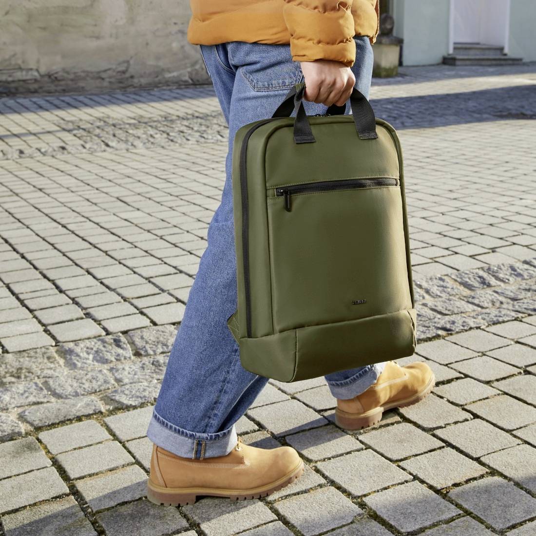 Person mit orangener Jacke und blauen Jeans hält grünen Rucksack, stehend auf gepflastertem Weg.