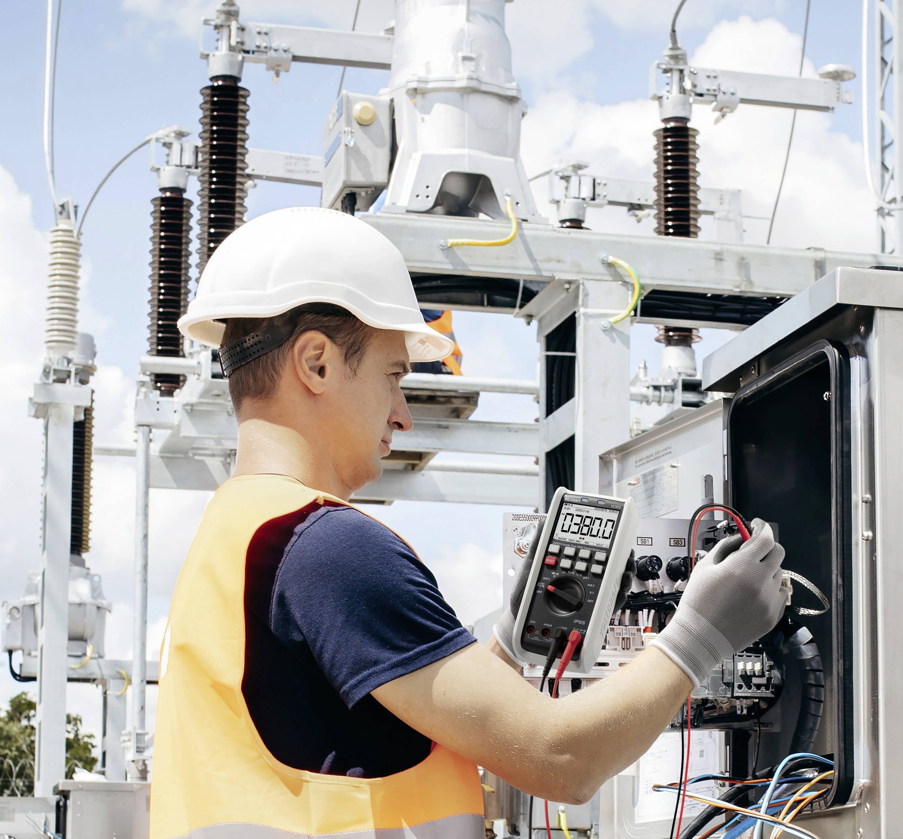 Techniker mit Schutzhelm und Sicherheitsweste verwendet ein Multimeter, um elektrische Ausrüstung in einer Freiluft-Transformatorenstation zu überprüfen.