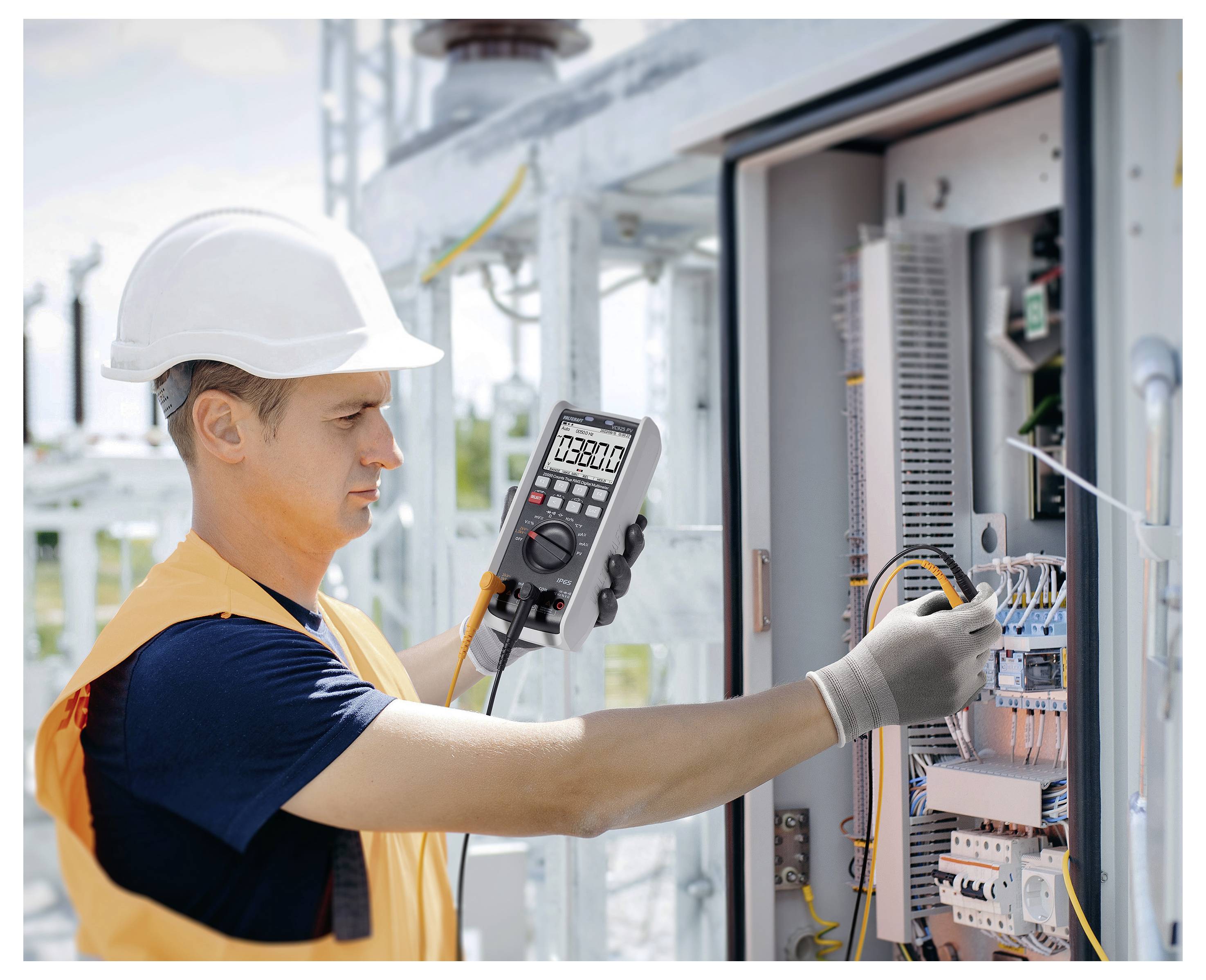 Elektriker mit Schutzhelm, der Verdrahtung in einem offenen elektrischen Schaltschrank mit einem digitalen Multimeter prüft.