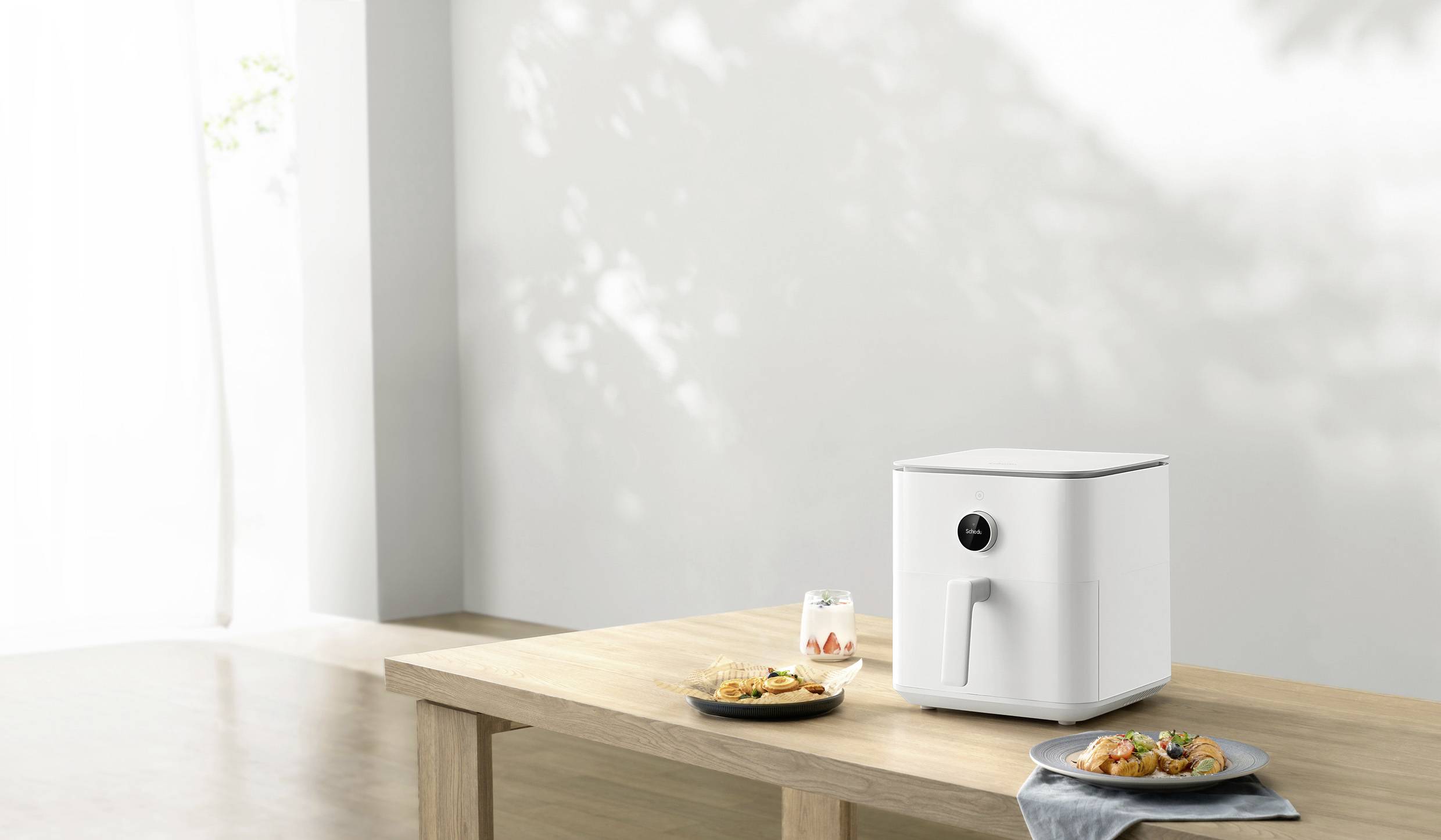 Ein weißer Airfryer steht auf einem hölzernen Tisch neben zwei Tellern mit zubereitetem Essen und einem Glas. Licht fällt durch ein Fenster.