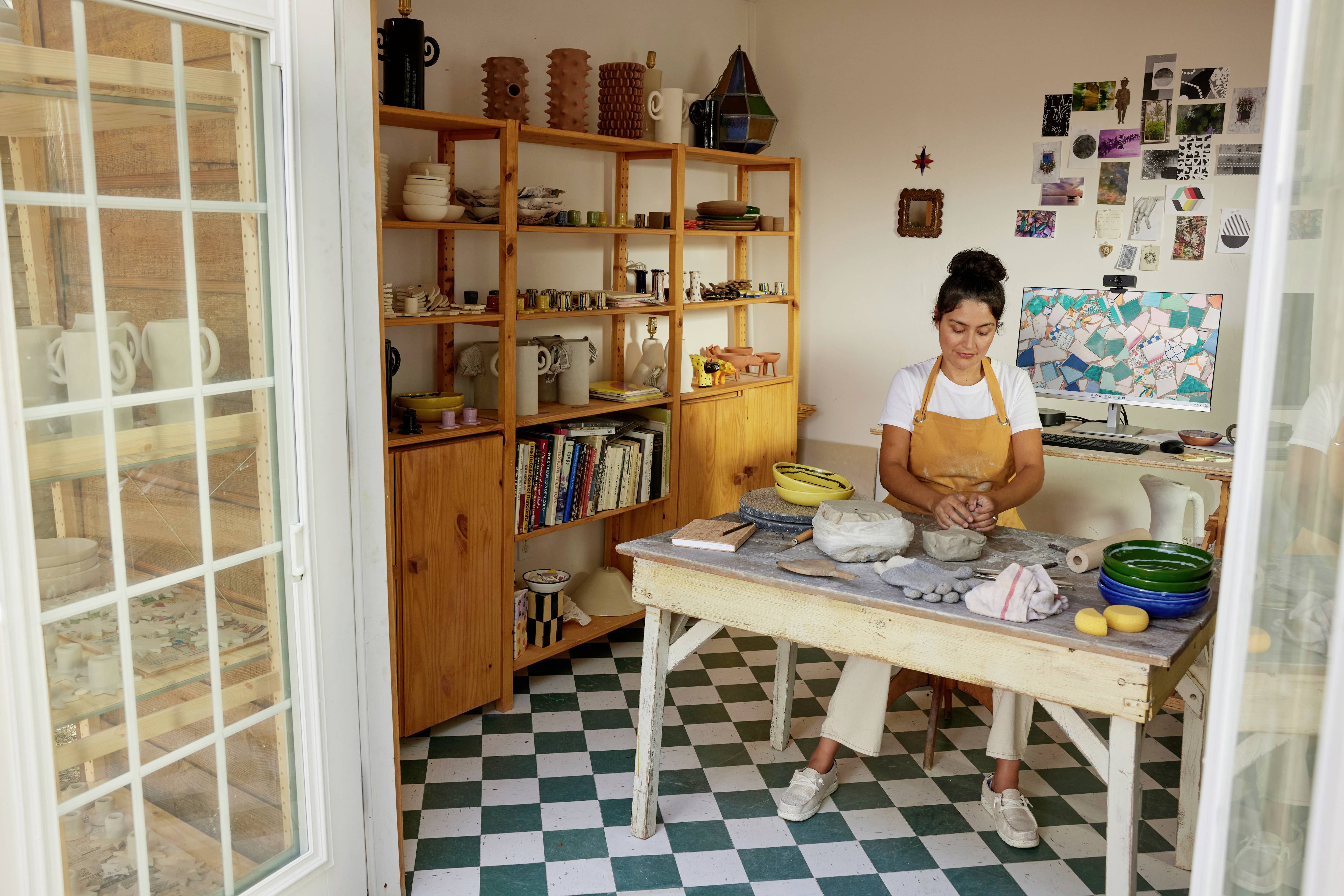 Eine Frau sitzt konzentriert an einem Tisch in einem hellen Keramikstudio. Sie formt Ton, umgeben von Keramikwerken und Regalen voller Materialien.