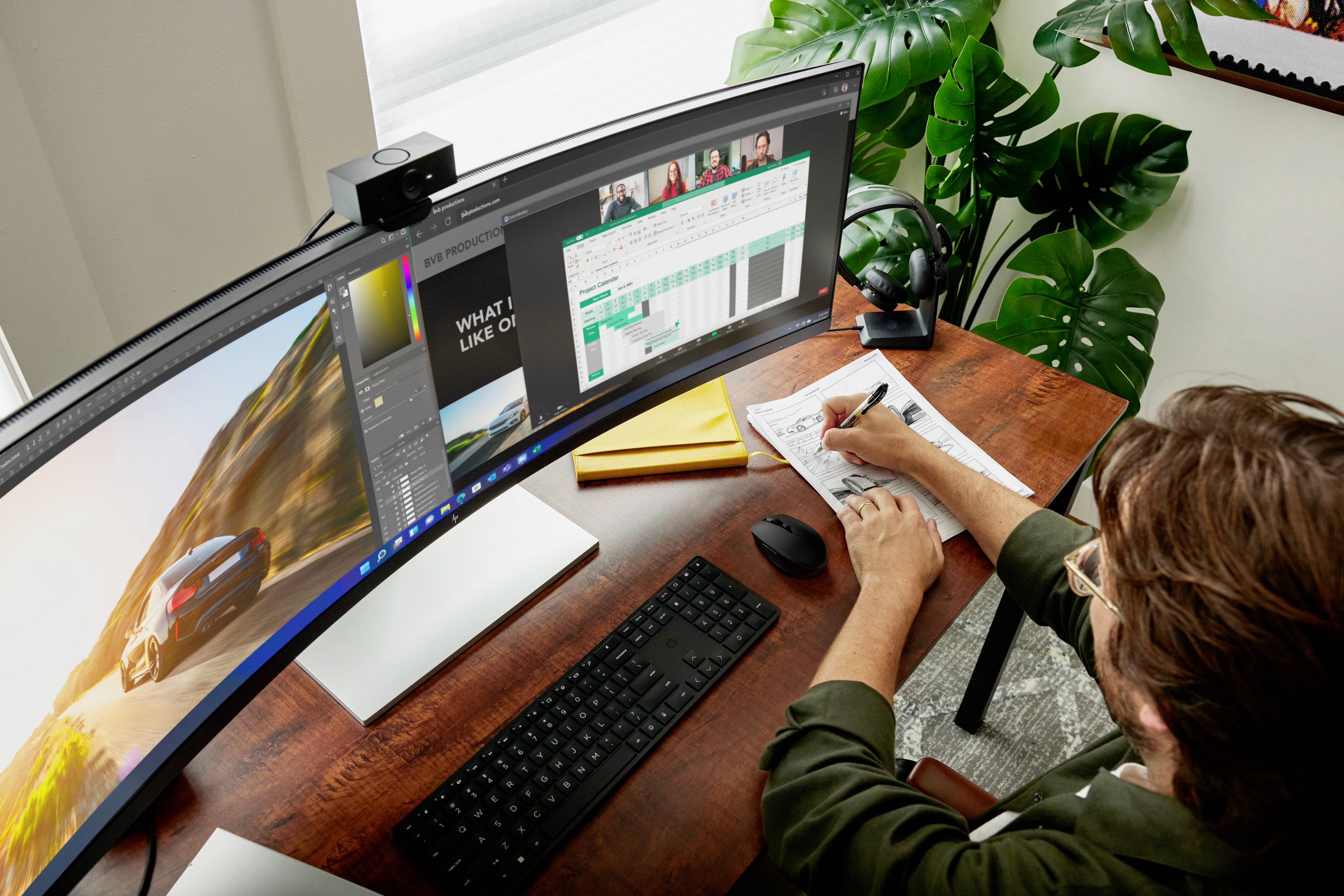 Person arbeitet an einem Schreibtisch mit einem großen, gebogenen Monitor, schreibt Notizen, während mehrere Programmfenster offen sind.