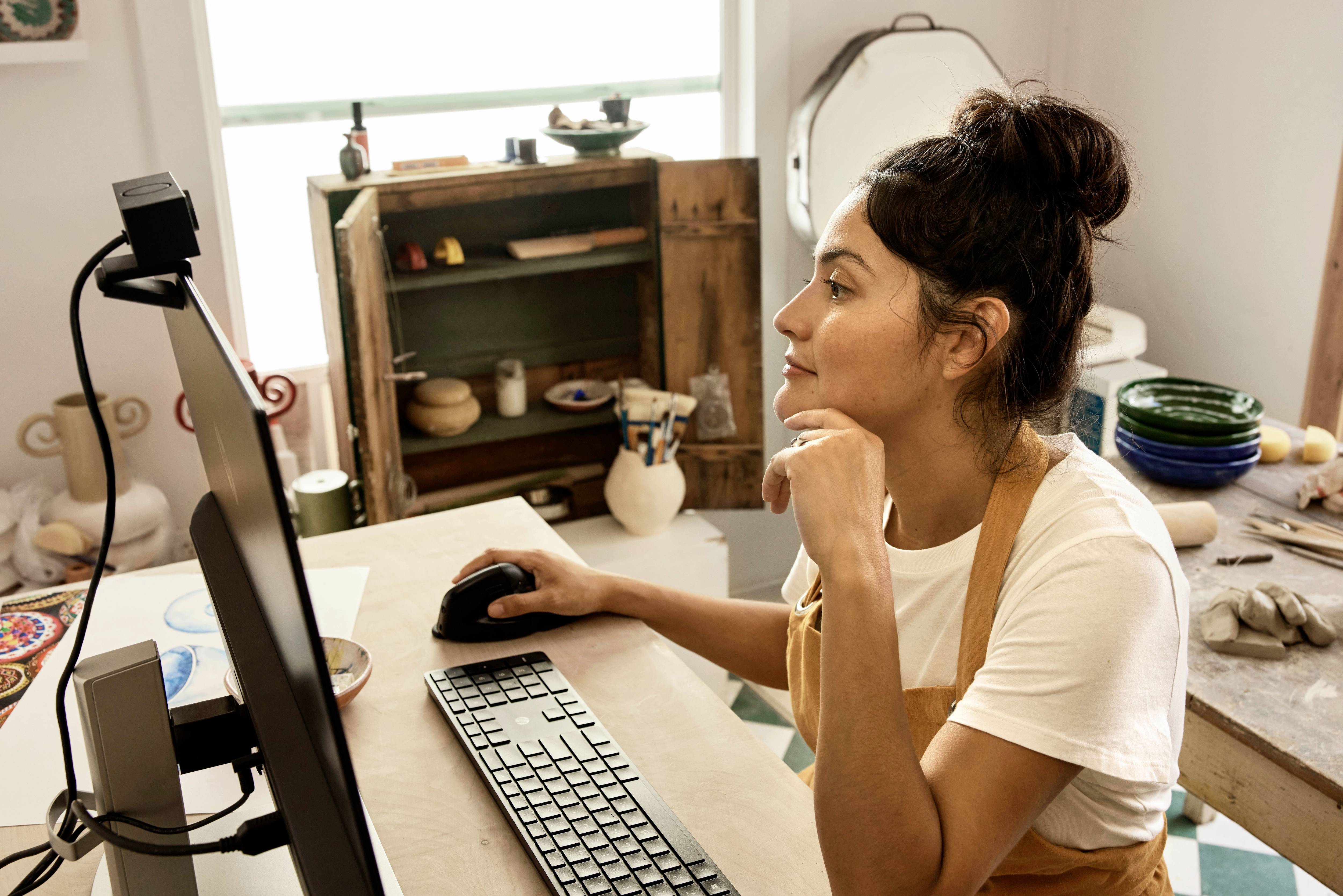 Eine Frau sitzt am Schreibtisch und schaut auf einen Computermonitor. Sie trägt ein weißes T-Shirt und eine Schürze. Im Hintergrund sind Töpferwaren zu sehen.