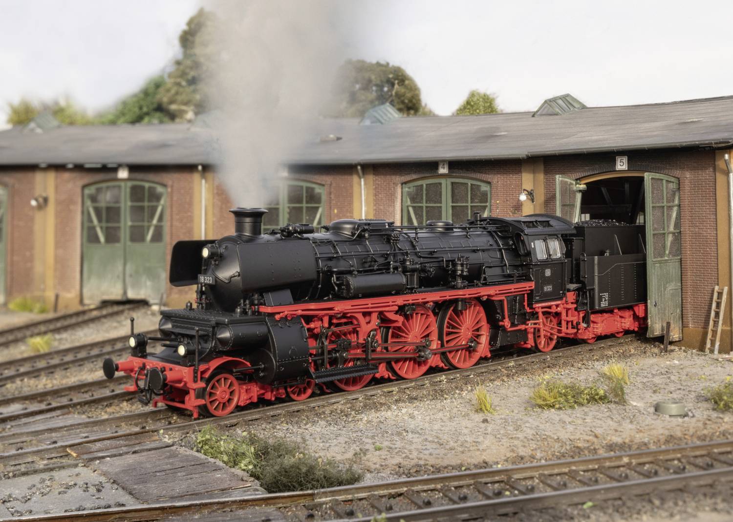 Eine schwarze Dampflokomotive mit roten Rädern steht vor einem Lokschuppen, aus dessen Schornstein Rauch aufsteigt.