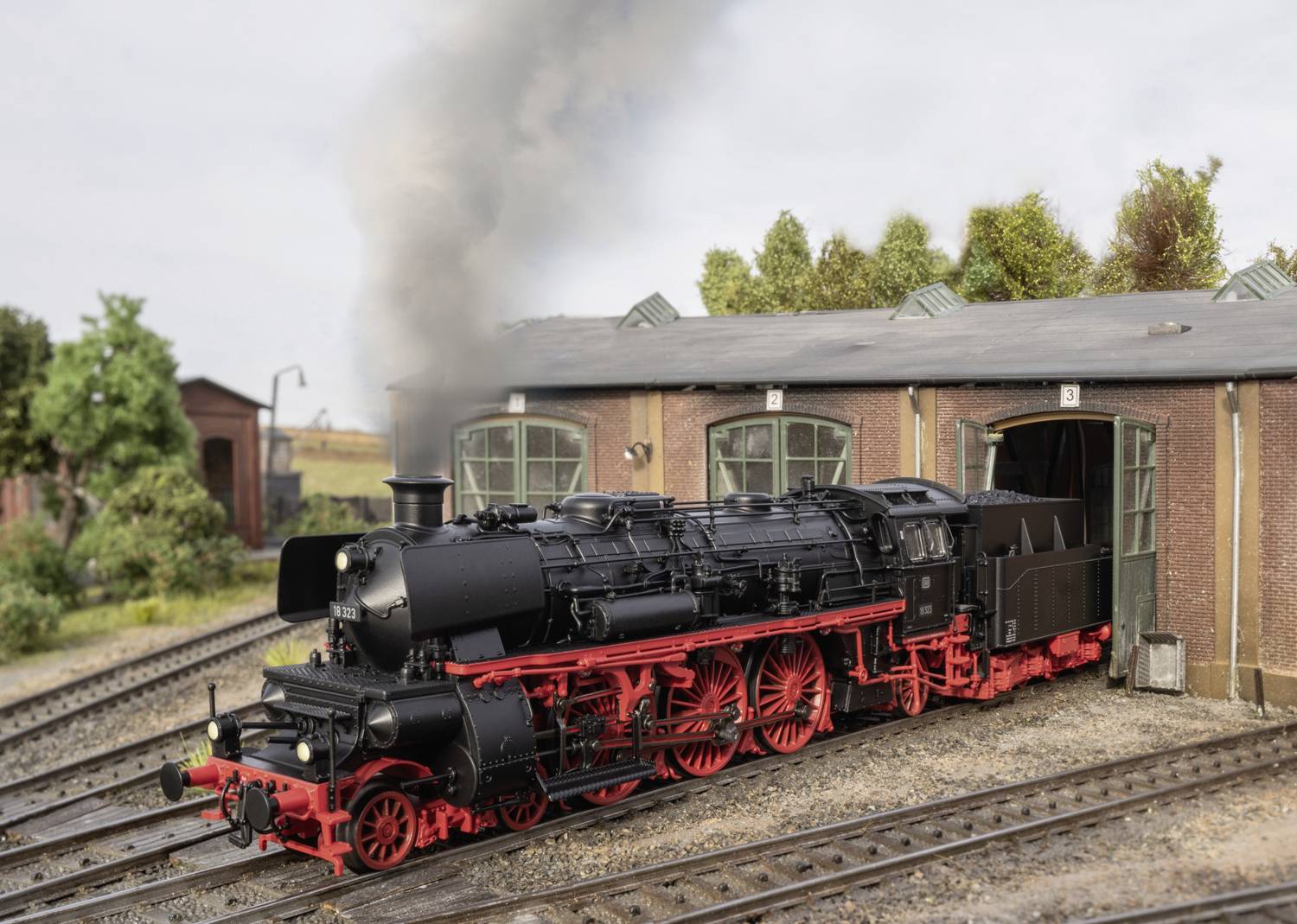 Eine schwarze Dampflokomotive mit roten Rädern fährt aus einem Bahndepot. Rauch steigt aus dem Schornstein auf. Im Hintergrund sind Bäume.