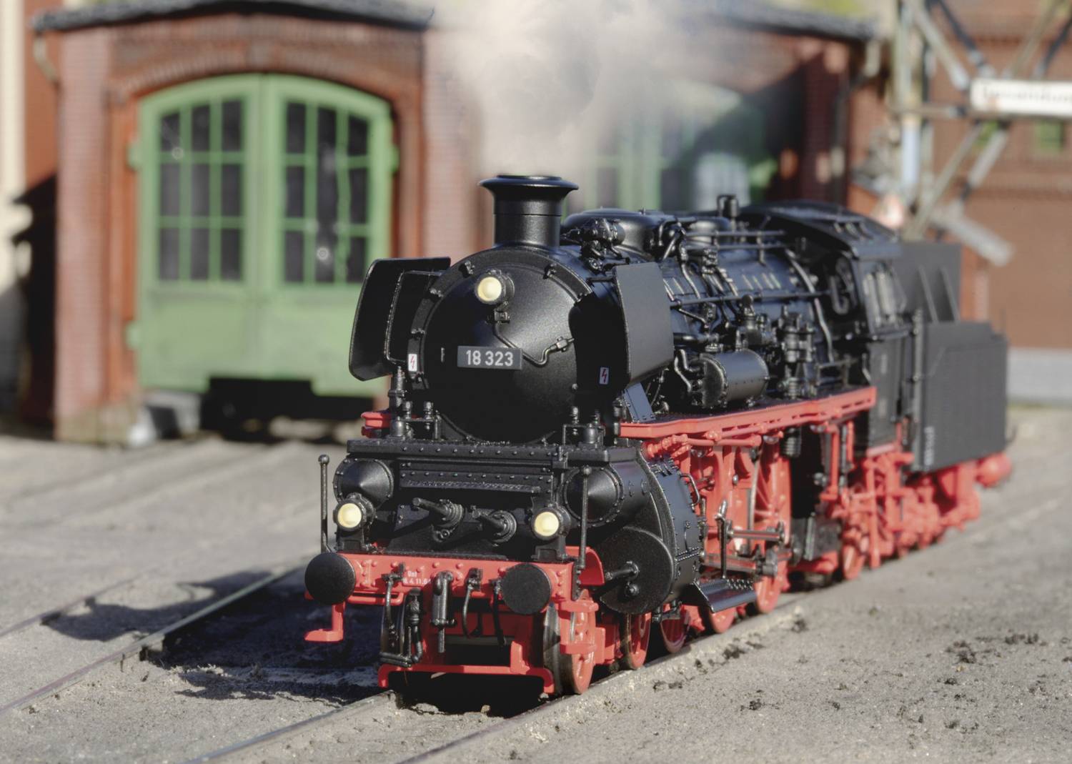 Eine schwarze Dampflokomotive mit rotem Fahrgestell fährt auf Schienen vor einem Gebäude mit grünen Toren. Rauch steigt aus dem Schornstein.
