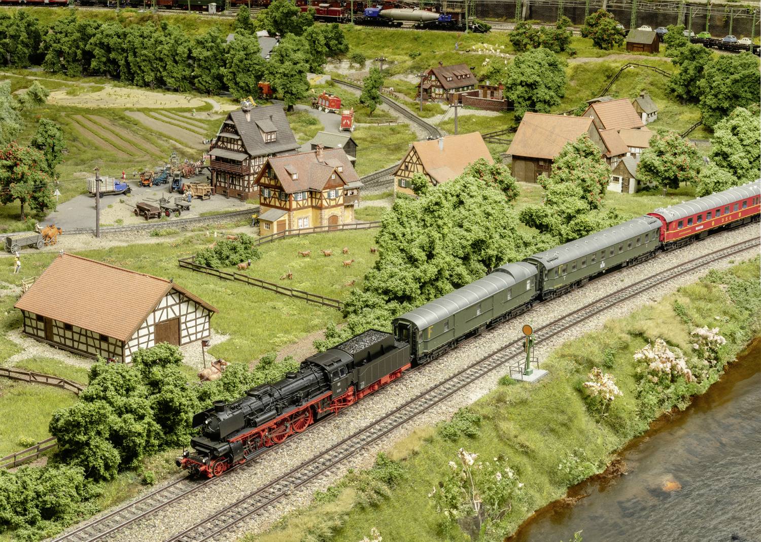 Eisenbahnmodell mit Landschaft: Ein schwarzer Zug fährt auf Schienen vorbei an Dörfern und Bäumen; detaillierte Miniaturszene.