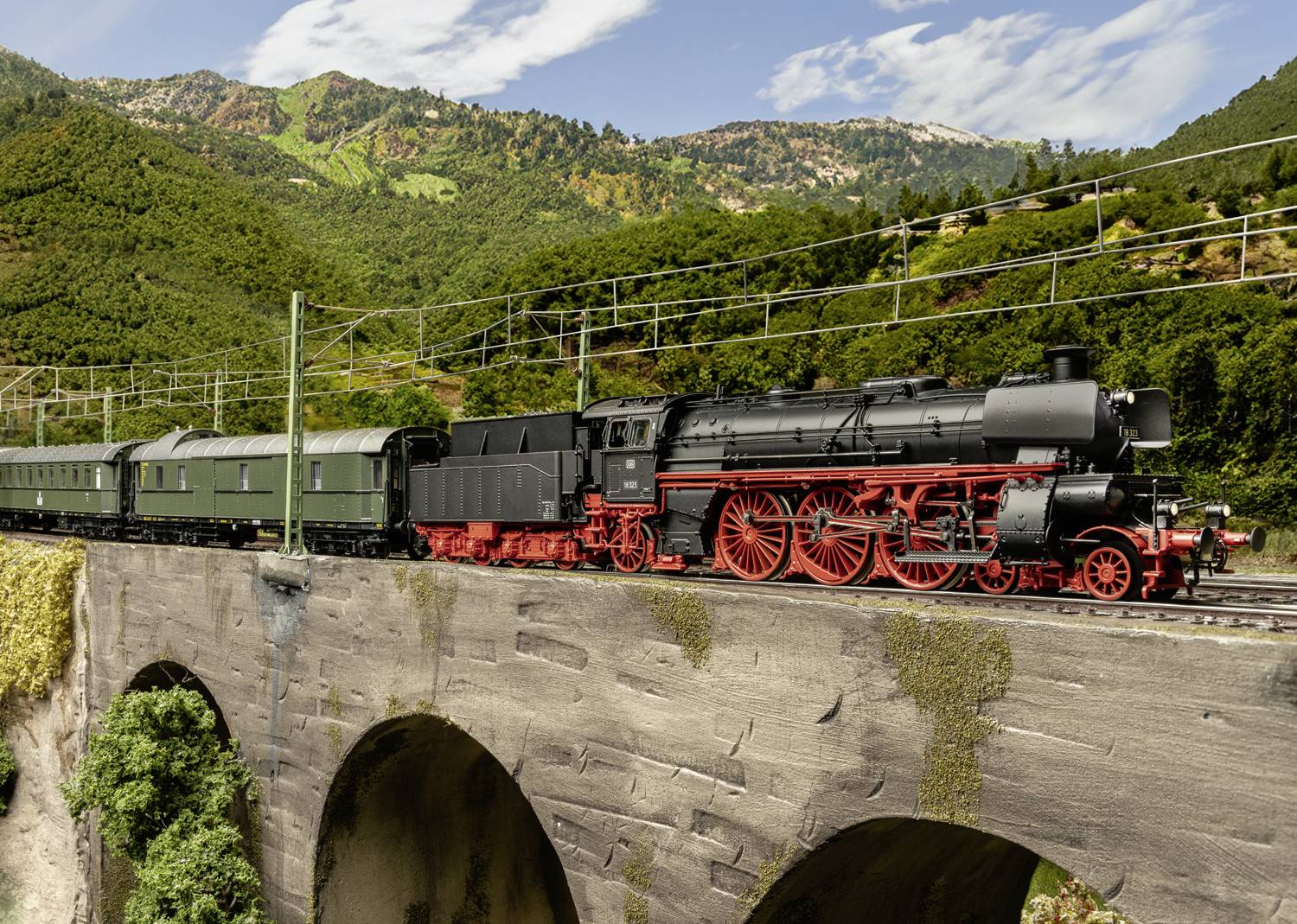 Eine schwarze Dampflokomotive mit roten Rädern zieht grüne Waggons über eine steinerne Brücke vor einer grünen Hügellandschaft.