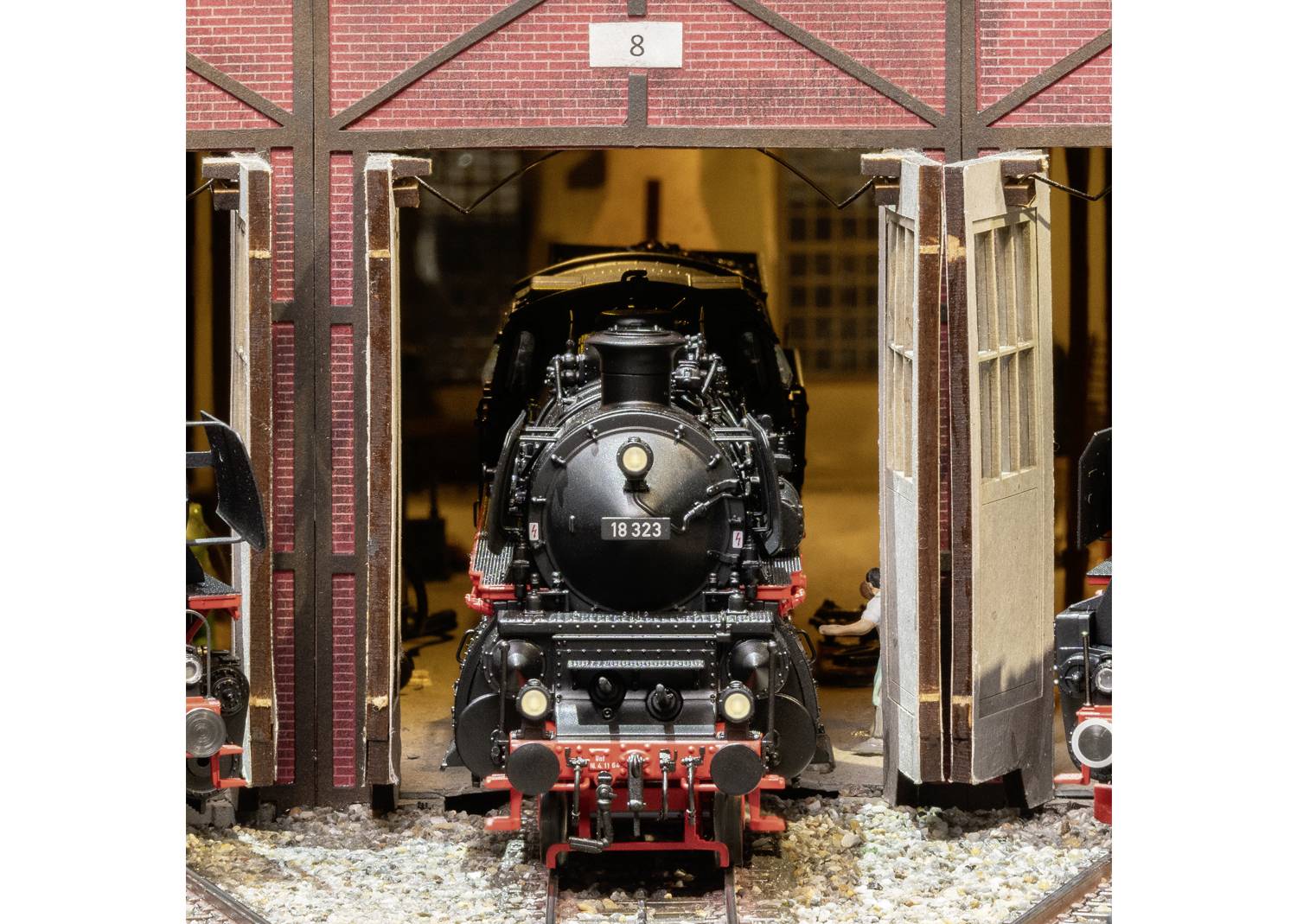 Eine schwarze Dampflokomotive mit der Nummer '18 323' steht in einem Backsteingebäude mit offener Tür. Oben eine '8' sichtbar.