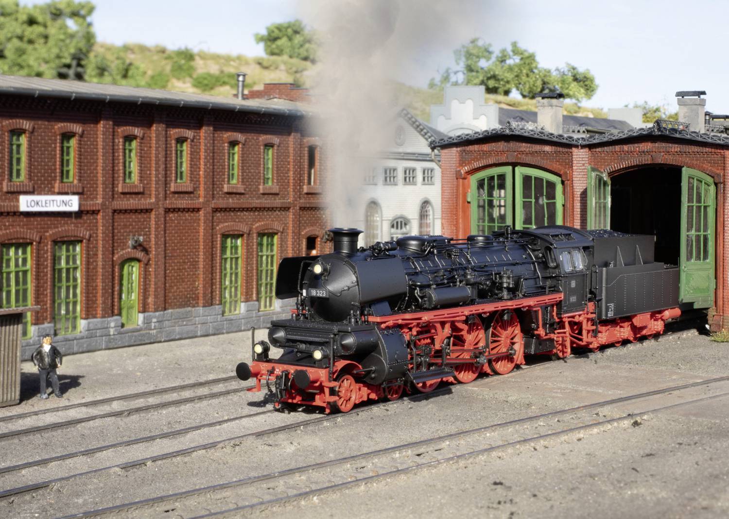 Eine schwarze Dampflokomotive steht auf Bahngleisen vor einem historischen Depot mit rotem Ziegelbau. Rauch steigt aus dem Schornstein.