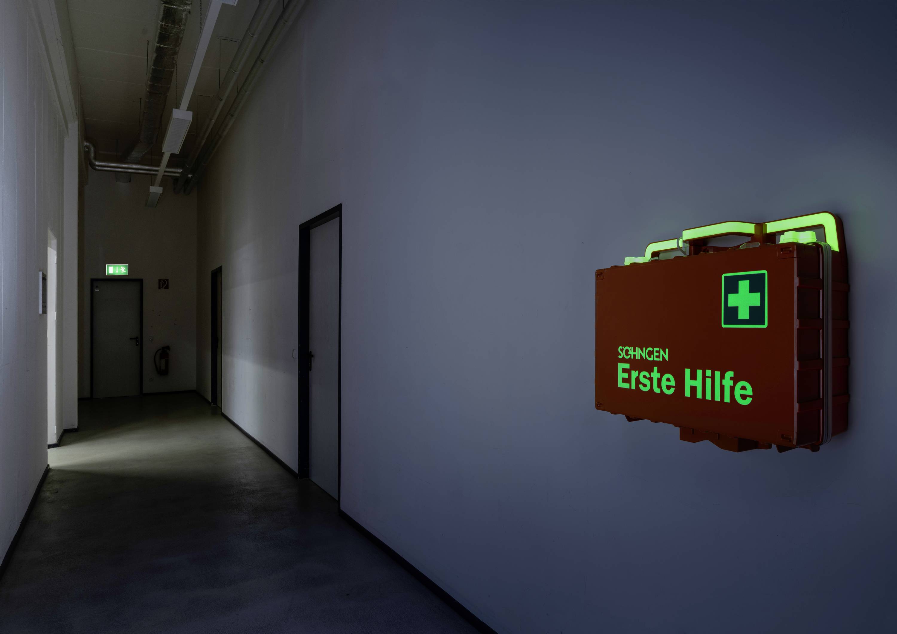 Erste-Hilfe-Koffer mit grünem Kreuz leuchtet im dunklen Flur an der Wand, über einer Tür ist ein Ausgangsschild zu sehen.