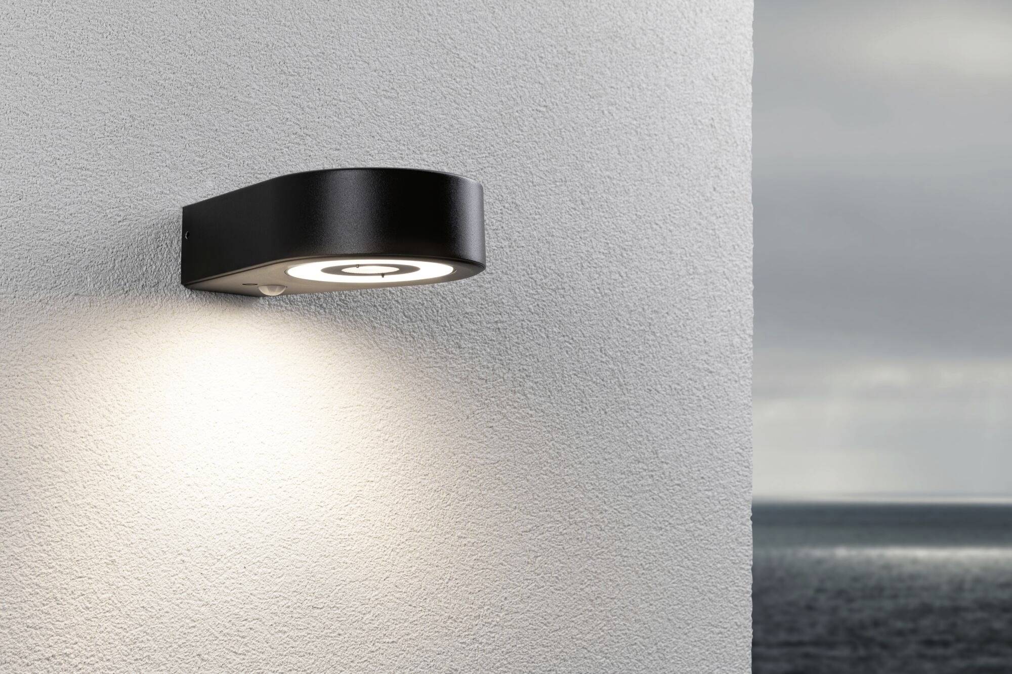 Eine moderne Wandaußenlampe leuchtet an einer weißen Wand; im Hintergrund ist das Meer bei bewölktem Himmel zu sehen.