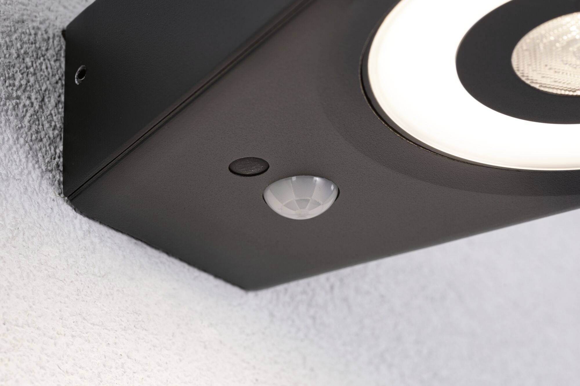 Eine schwarze Außenlampe mit Bewegungssensor ist an einer weißen Wand befestigt. Sie hat eine runde Leuchte an der Unterseite.