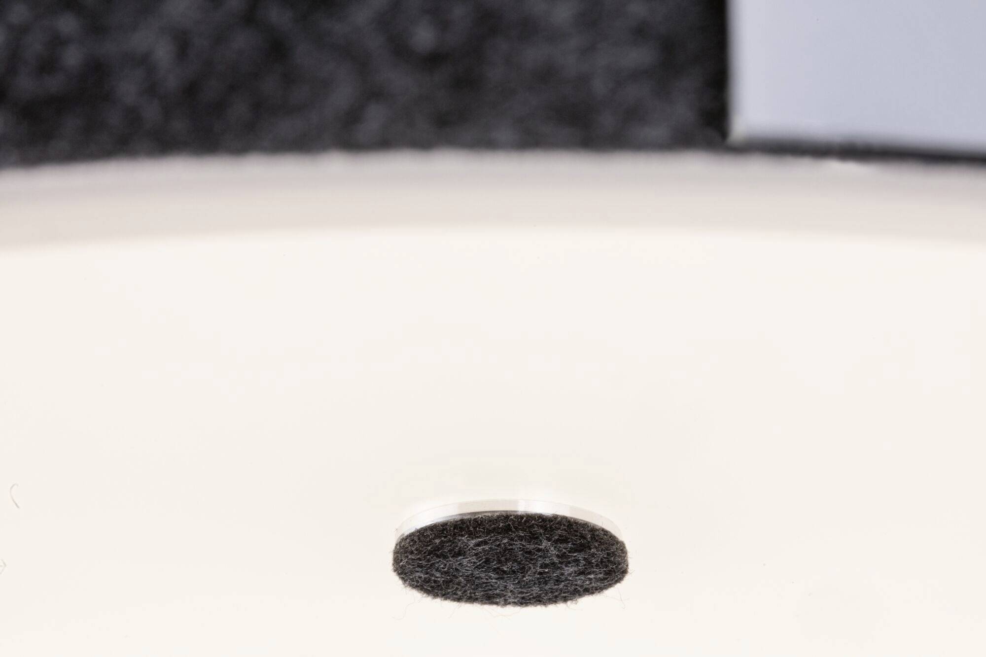 Ein runder, dunkler Filzgleiter auf einer glatten, weißen Oberfläche. Links oben ist ein unscharfer, dunkler Hintergrund sichtbar.