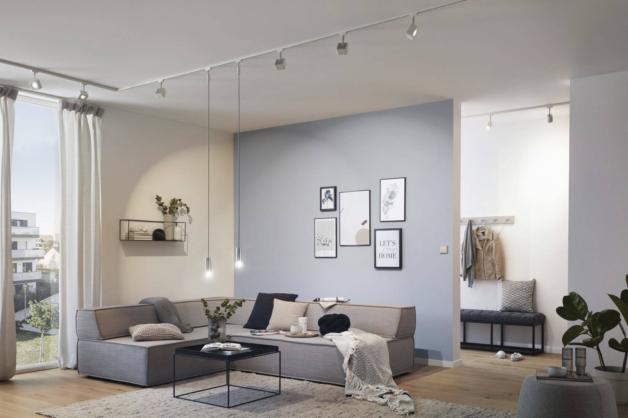 Ein modernes Wohnzimmer mit Ecksofa, Couchtisch und dekorativer Wand mit Bilderrahmen. Durch das Fenster fällt Tageslicht ein.