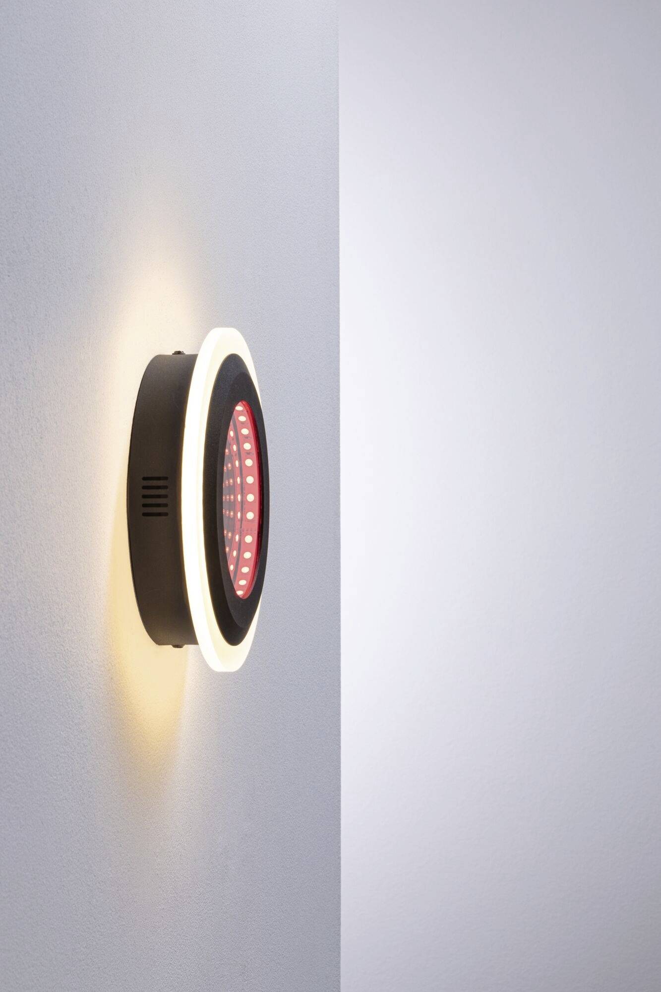 Wandleuchte mit schwarzem Gehäuse, rotem LED-Lichtzentrum und weißem Leuchtring, auf eine weiße Wand montiert.