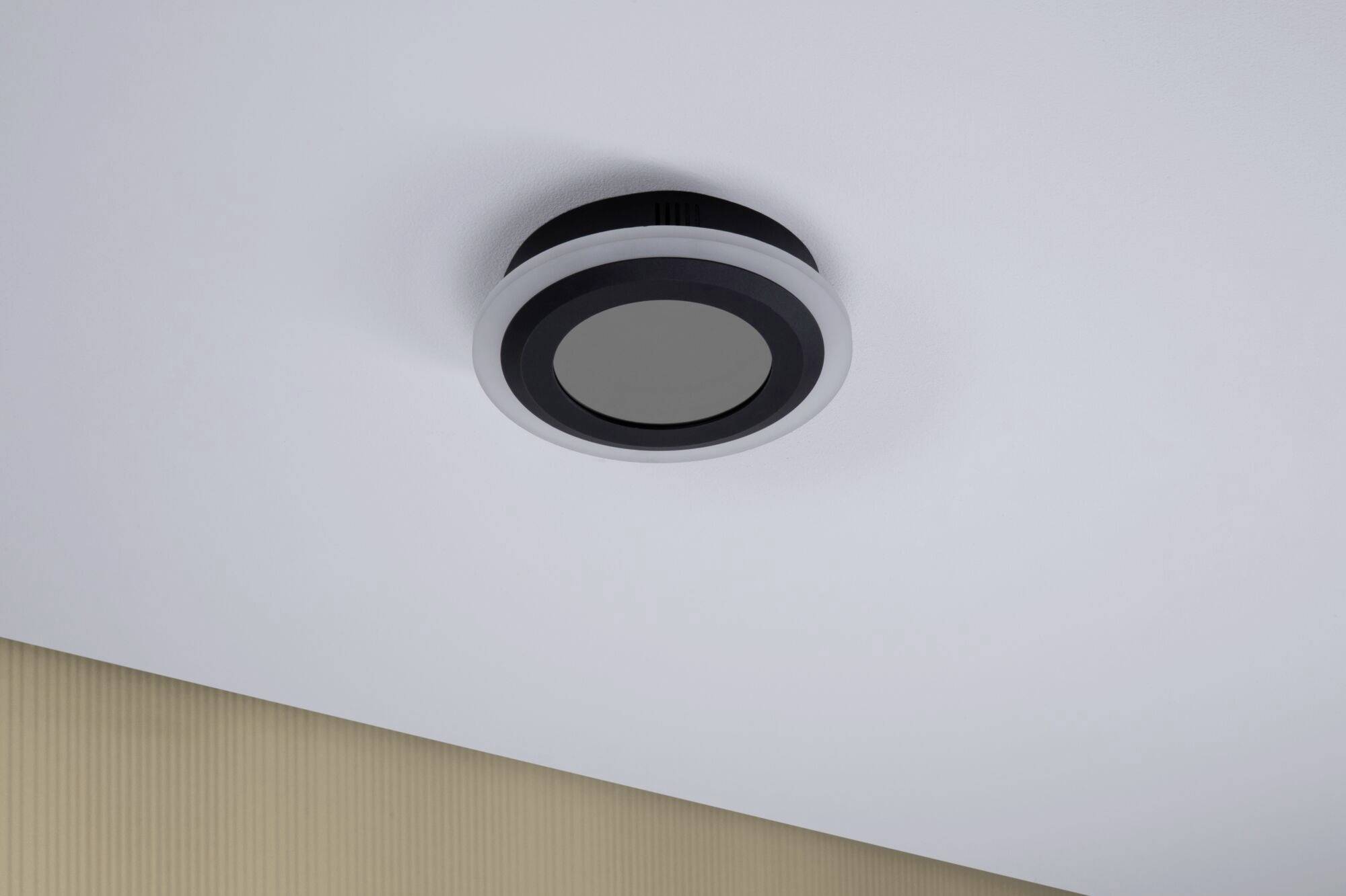 Ein runder Rauchmelder ist an einer weißen Decke montiert. Die Wand darunter ist beige. Das Gerät ist schlicht in Schwarz und Weiß gehalten.