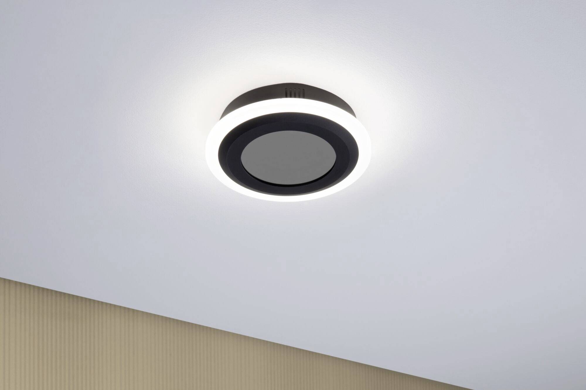 Schwarze, runde Deckenleuchte mit leuchtendem LED-Ring, an weißer Decke montiert über einer beigen Wand. Modernes Design.