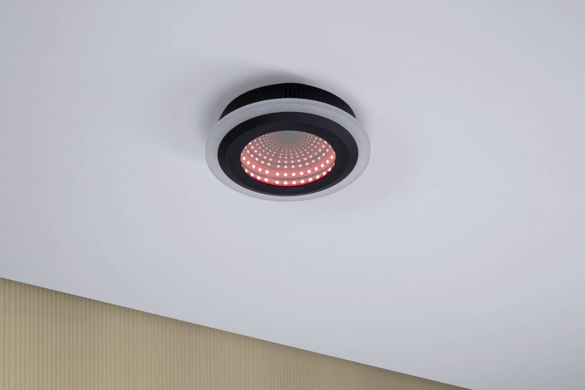 Eine schwarz-graue Deckenleuchte mit rotem LED-Licht ist an einer weißen Decke montiert. Sie leuchtet in einem modernen Raum.