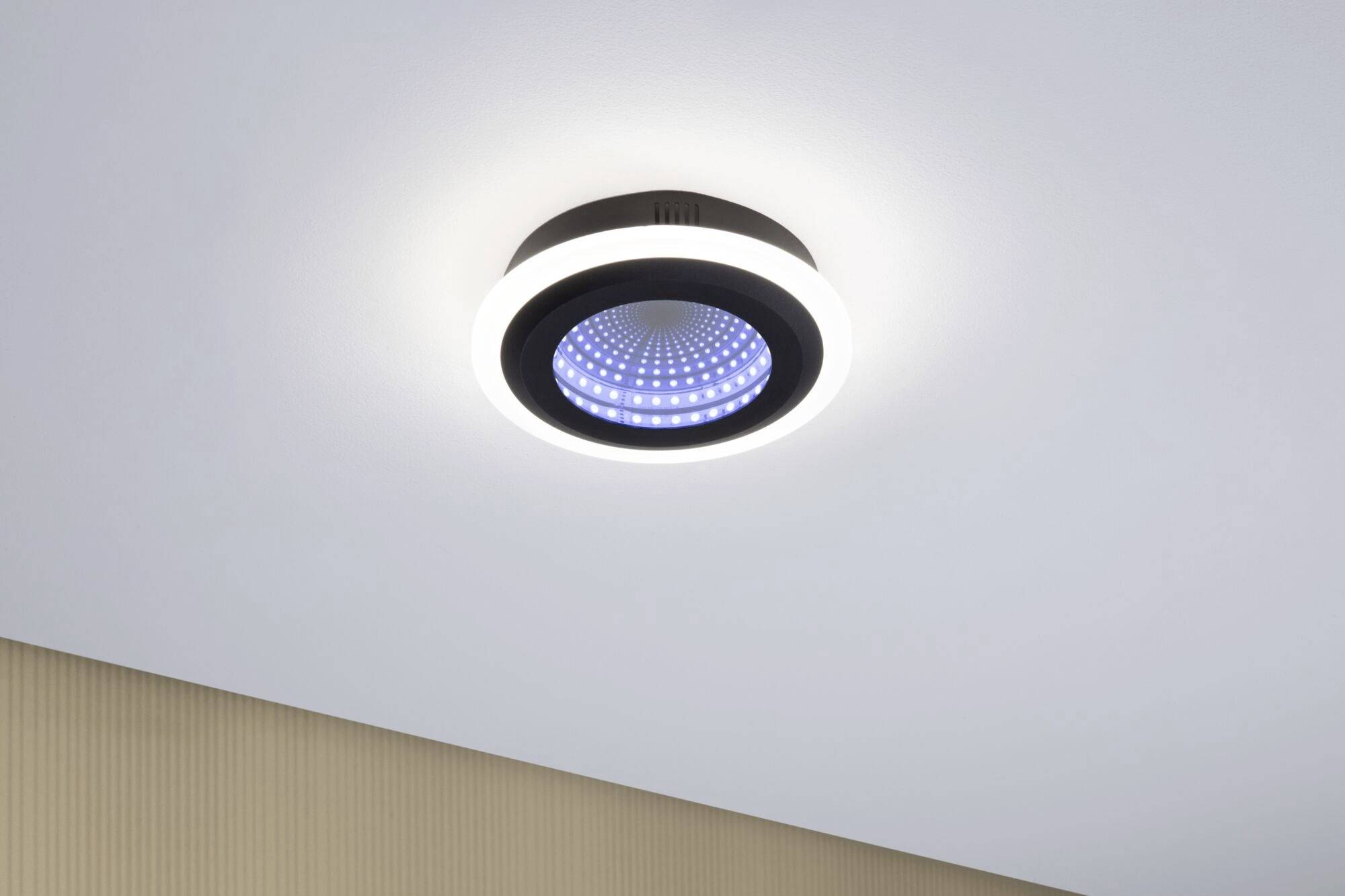 Eine moderne Deckenleuchte strahlt blaues und weißes Licht aus und ist an einer weißen Decke in einem Raum mit beige Wand montiert.