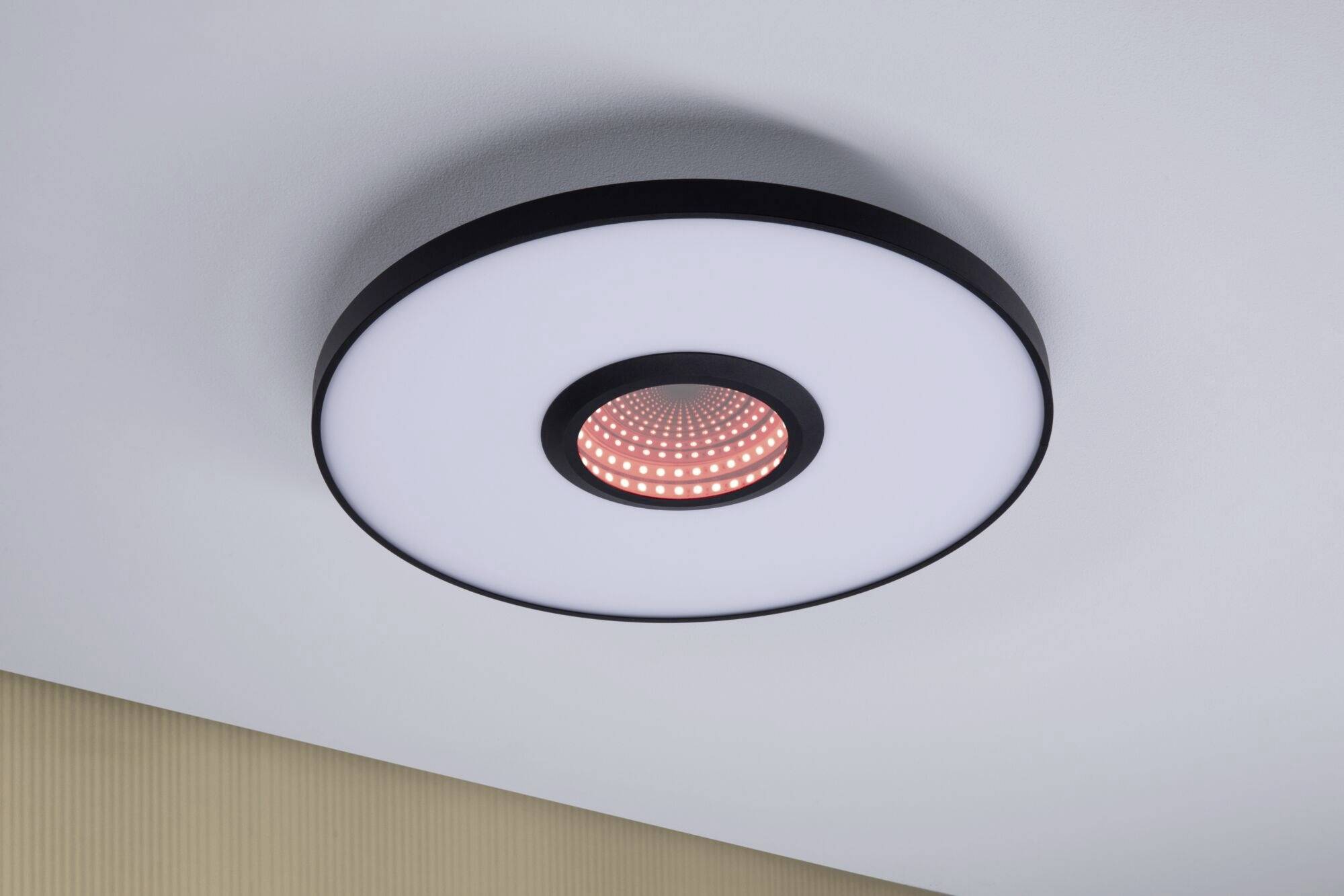 Runde Deckenleuchte mit LED, leuchtet zentral rot. Befestigt an einer weißen Decke über einem beige gestreiften Wandabschnitt.