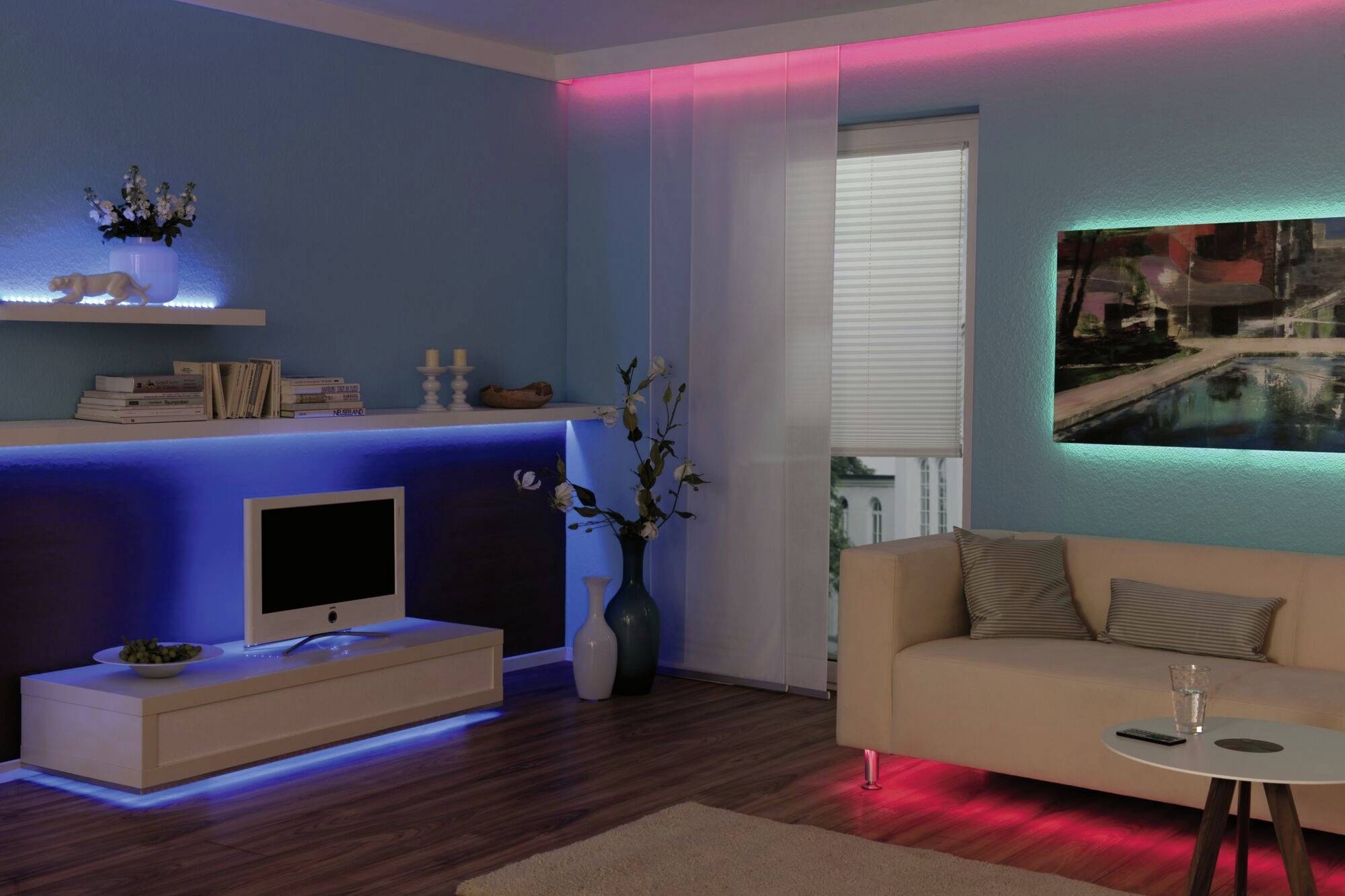 Beleuchtetes Wohnzimmer mit blauen und roten LED-Leuchten an Decke und Möbeln. Moderne Möbel, Vase mit Blumen, Bild an der Wand.