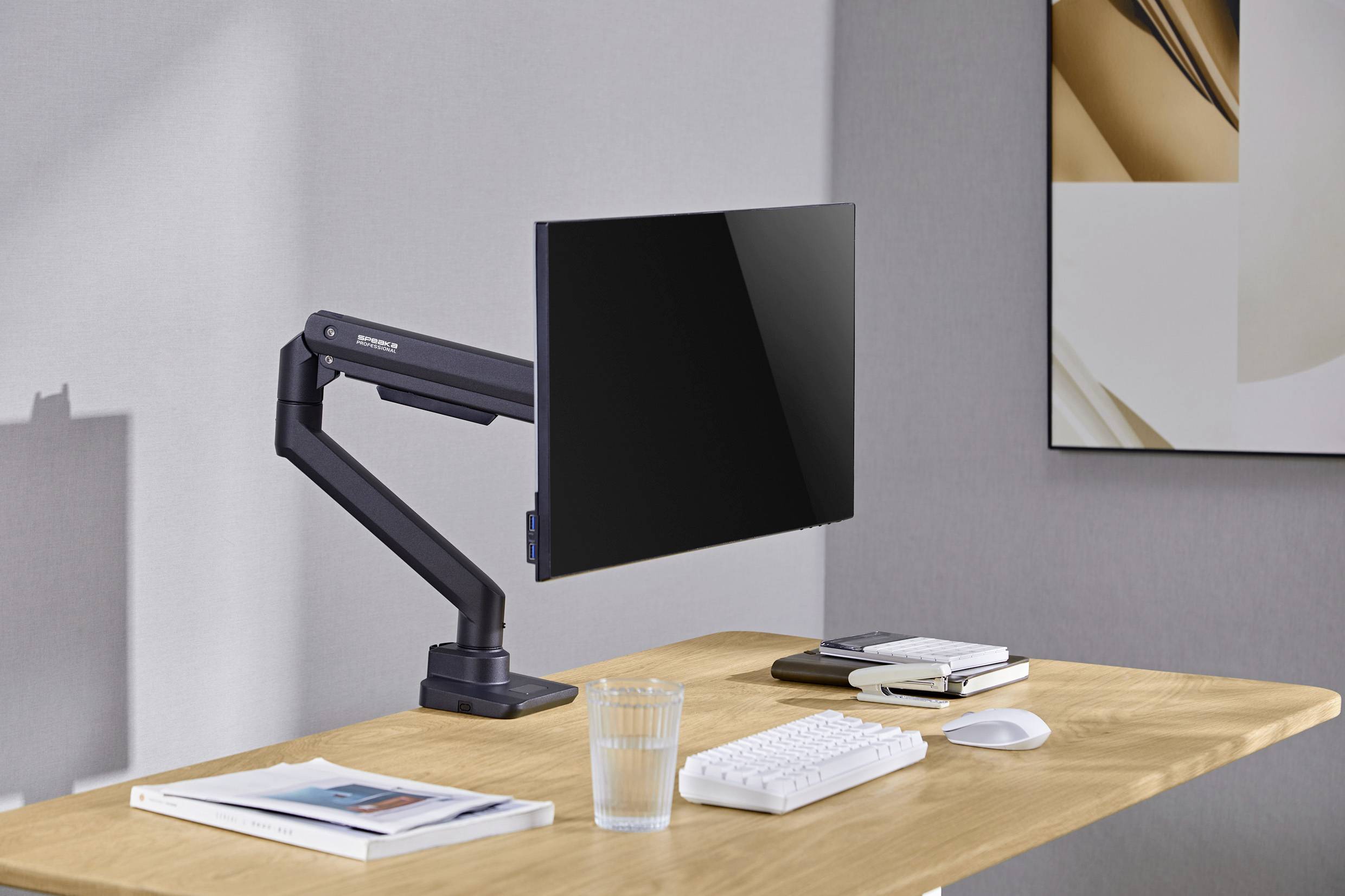 Ein Schreibtisch aus Holz mit einem schwarzen Monitor an einem verstellbaren Arm. Darauf eine Tastatur, Maus, Glas Wasser und zwei Bücher.