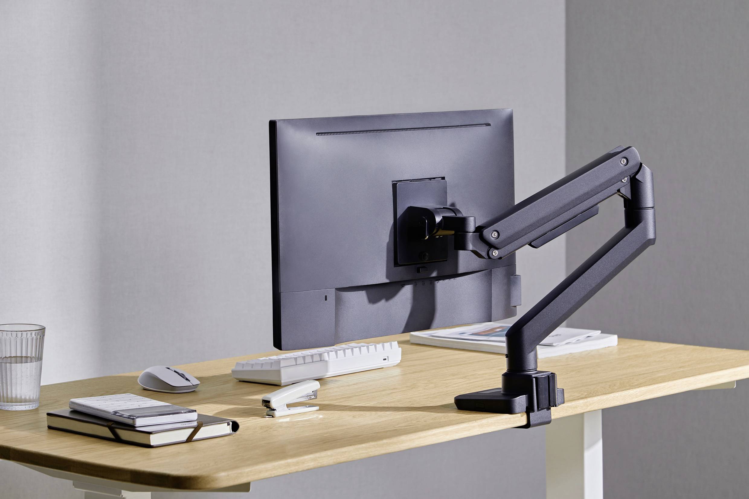 Ein minimalistischer Arbeitsplatz mit Monitor auf einem verstellbaren Arm, daneben Tastatur, Maus, Notizbuchstapel und Glas Wasser.