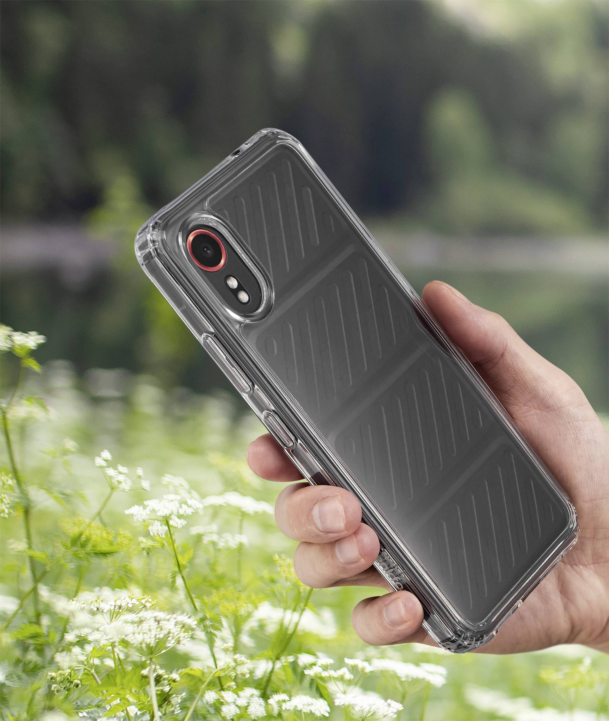 Eine Hand hält ein Smartphone mit einer durchsichtigen Hülle vor einer unscharfen, grünen Waldlandschaft mit einem See im Hintergrund.