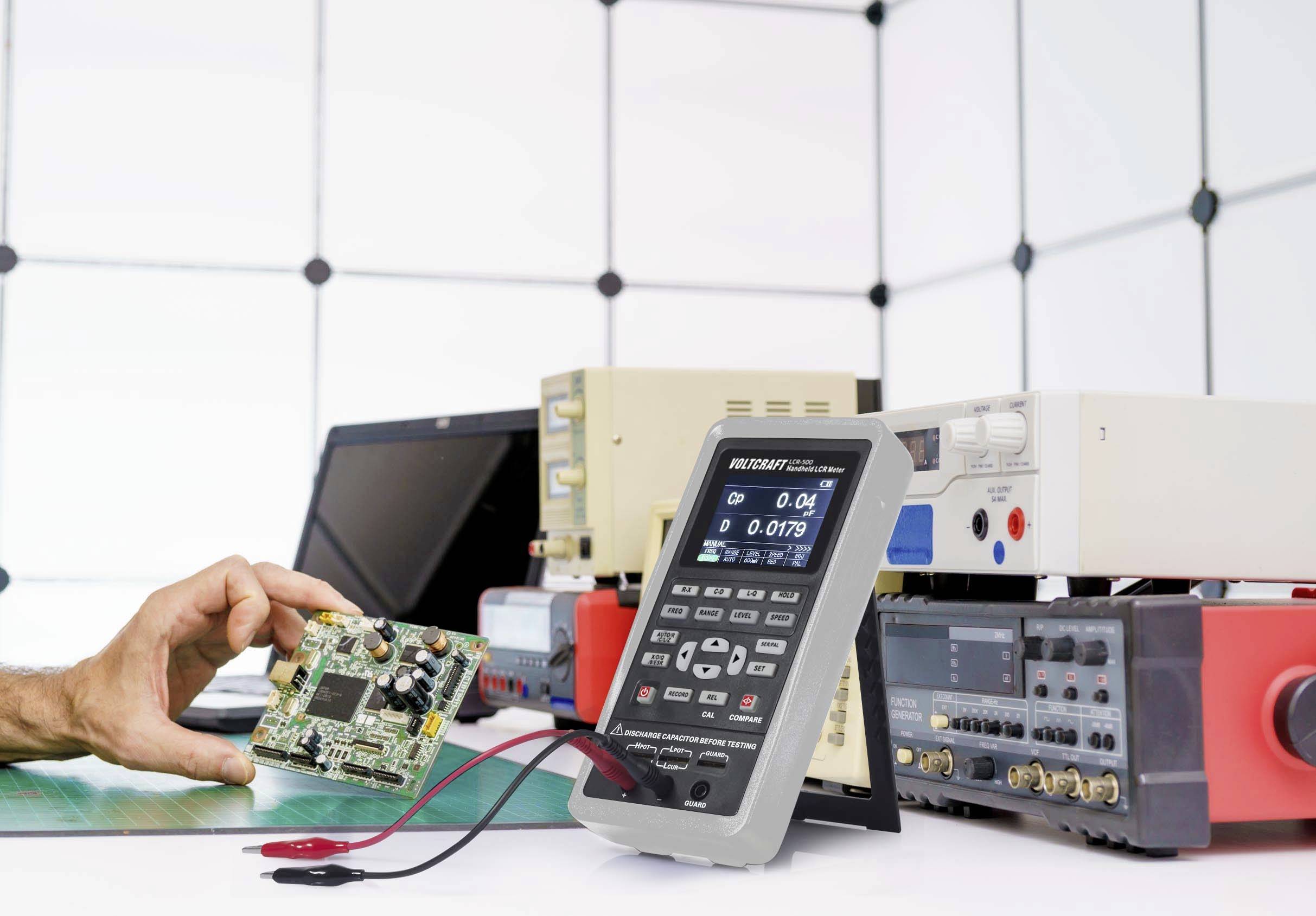 Eine Person hält eine Computerplatine neben mehreren elektronischen Messgeräten auf einem Tisch. Es handelt sich um ein Laborumfeld.