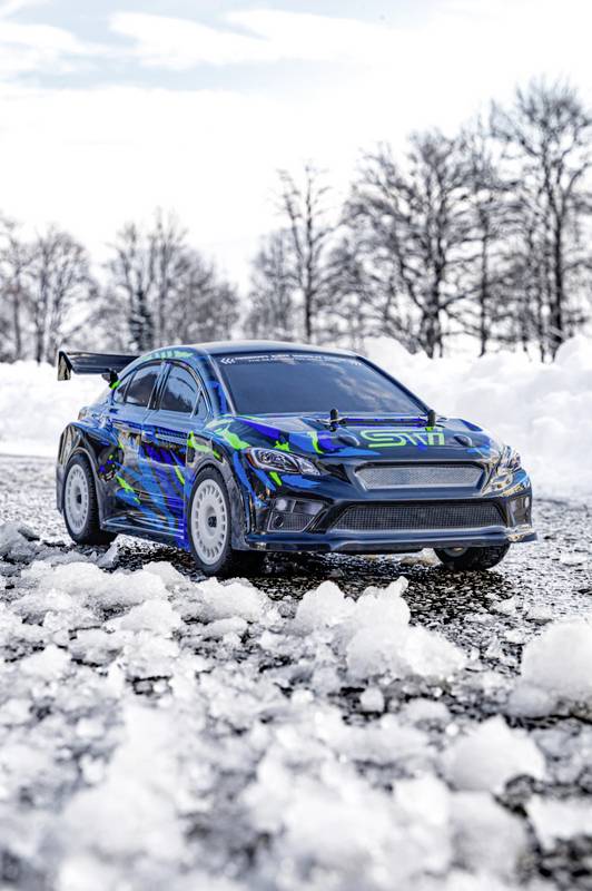 Ein Rallyeauto mit blauer und grüner Lackierung fährt durch eine schneebedeckte Landschaft. Im Hintergrund sind verschneite Bäume.