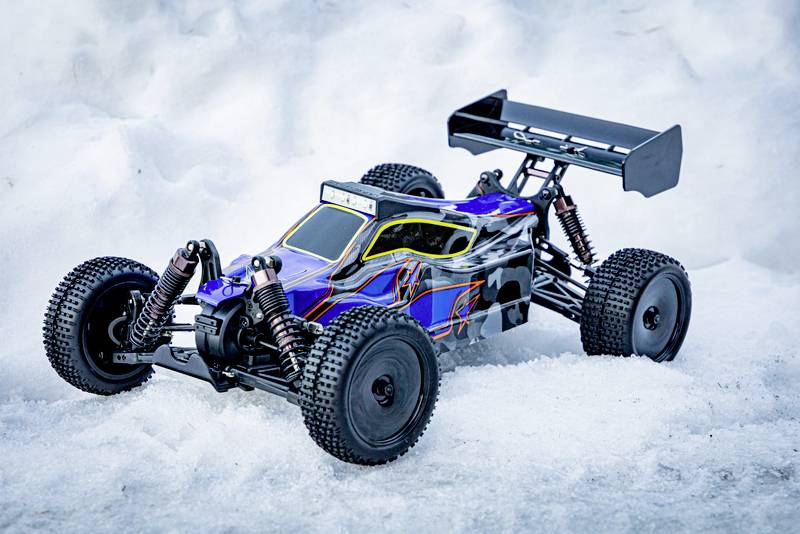 Ein blau-gelbes ferngesteuertes Auto mit schwarzen Rädern steht im Schnee. Es hat eine große Heckflügel für Stabilität.