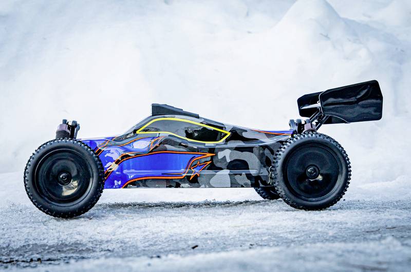 Ein ferngesteuertes Auto mit blauem und schwarzem, camouflagemusterbemaltem Chassis, steht auf schneebedecktem Boden. Es hat große Reifen.