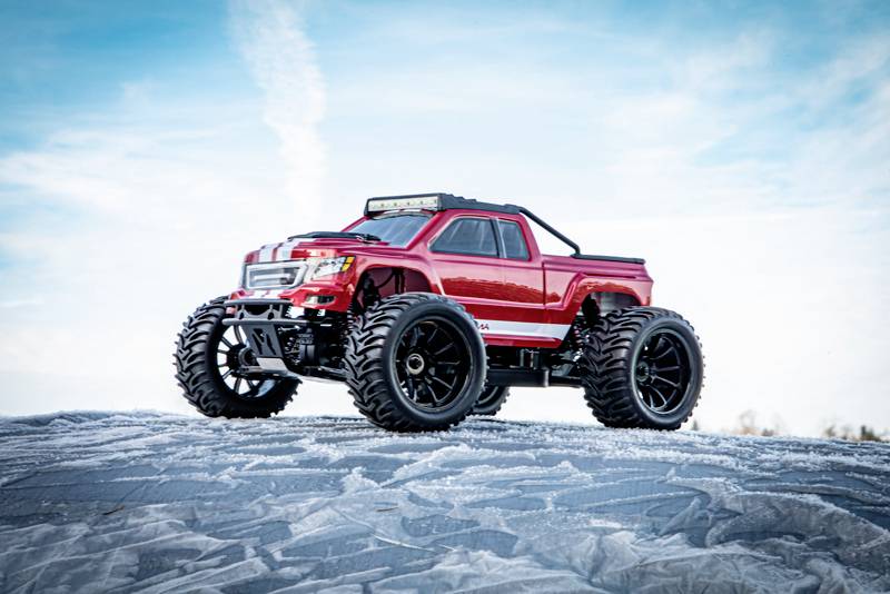 Ein rotes ferngesteuertes Auto mit großen Offroad-Reifen steht auf schneebedecktem Untergrund vor einem blauen Himmel.