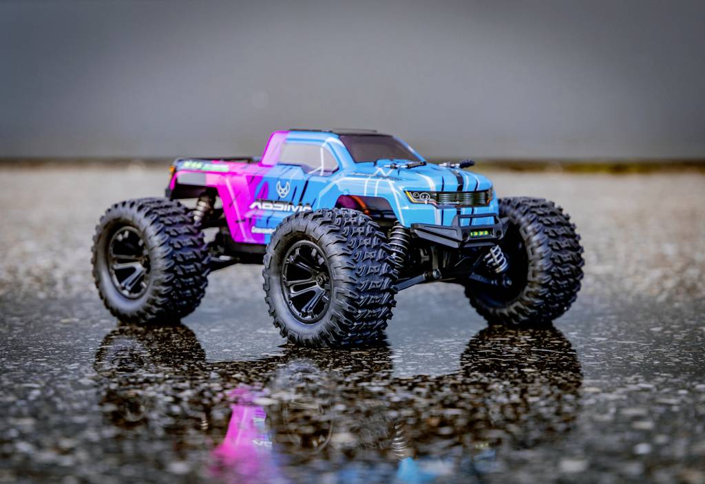 Spielzeug-Monstertruck in Blau und Pink auf nassem Asphalt reflektiert. Grobe Reifen, moderne Karosserie, dynamisches Design.