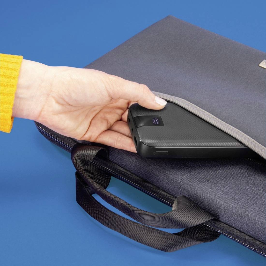 Eine Hand zieht ein schwarzes Smartphone aus einer schwarzen Tasche auf einem blauen Untergrund.