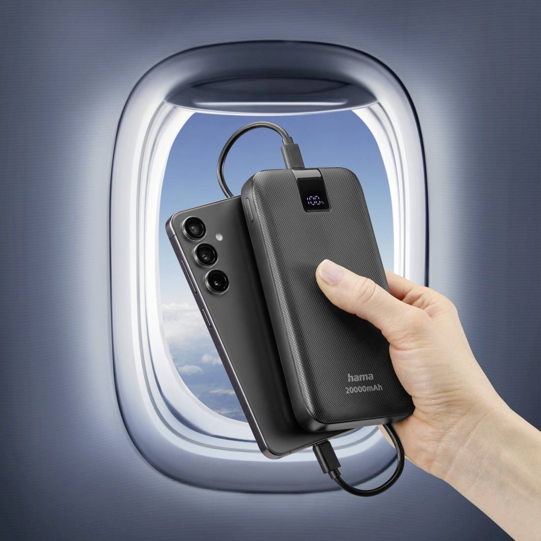 Eine Hand hält eine Powerbank und ein Smartphone vor einem Flugzeugfenster. Beide Geräte sind über ein Kabel verbunden.