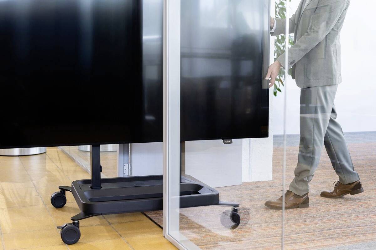 Person in einem Büroanzug schiebt einen großen Monitor auf Rollen durch einen Glasdurchgang. Im Hintergrund sind Pflanzen sichtbar.