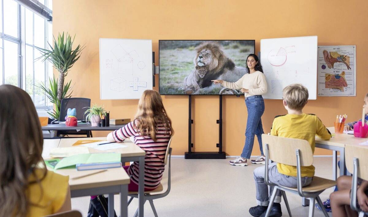 Lehrerin zeigt in einem Klassenzimmer auf einen Bildschirm mit Löwenbild. Kinder hören aufmerksam zu. Schulisches Lernen in natürlicher Umgebung.