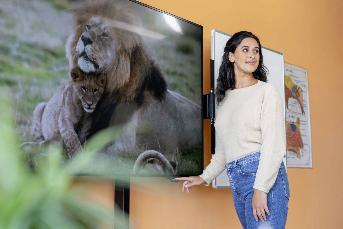 Eine Frau steht neben einem Bildschirm, auf dem ein Löwe mit einem Jungtier gezeigt wird. Im Hintergrund sind wissenschaftliche Poster.